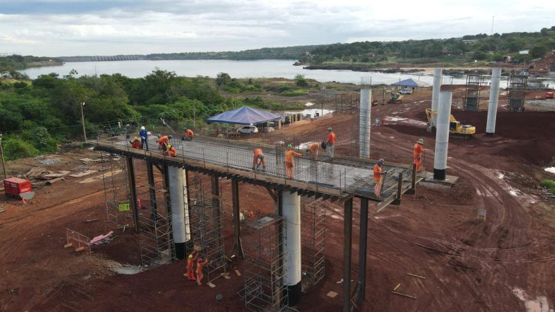 Construction workers in orange safety gear assemble an elevated steel walkway on concrete columns above a muddy site beside a river with green hills in the background and tents nearby.